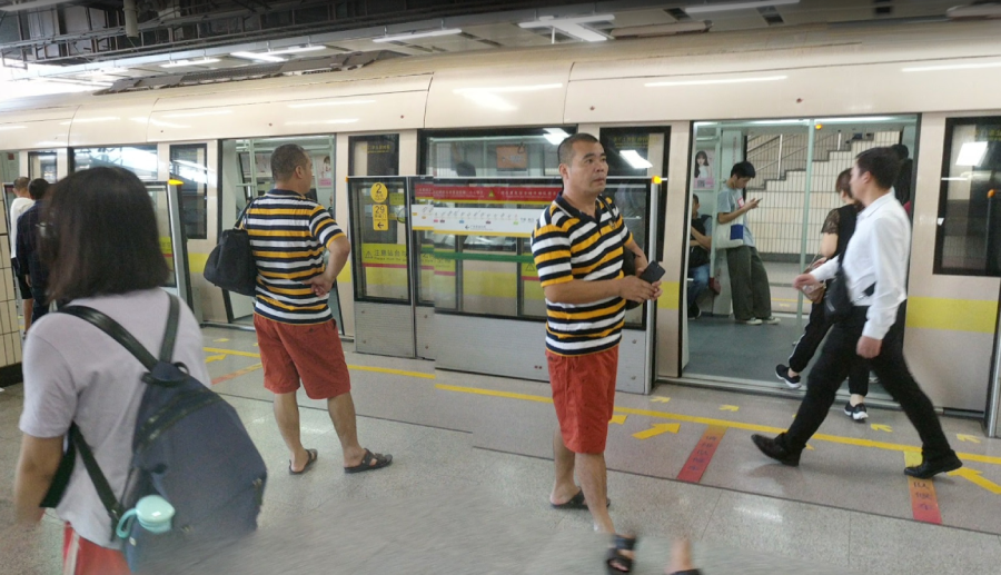A man in a yellow, black, and white striped shirt with orange shorts and sandals appears twice on a subway platform due to a photo editing effect. Other commuters are nearby, some boarding the train.