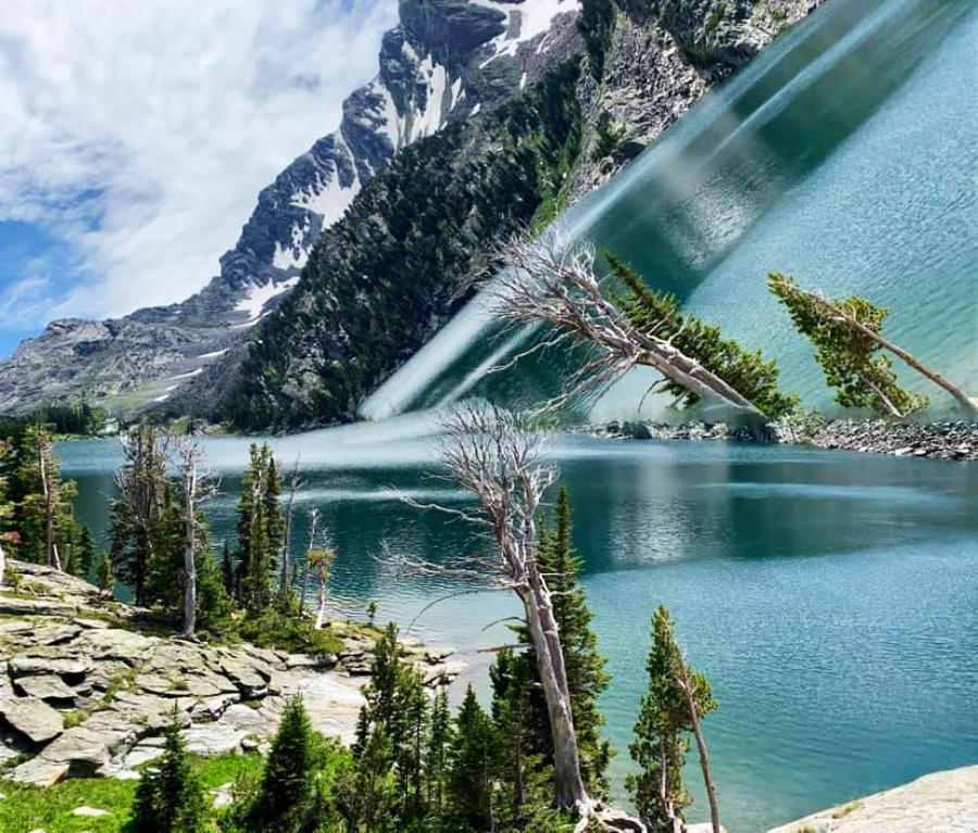 A serene mountain lake scene is distorted by a diagonal split, bending trees and water unnaturally, with snow-capped peaks, rocky shore, and pine trees creating a surreal mirrored effect.