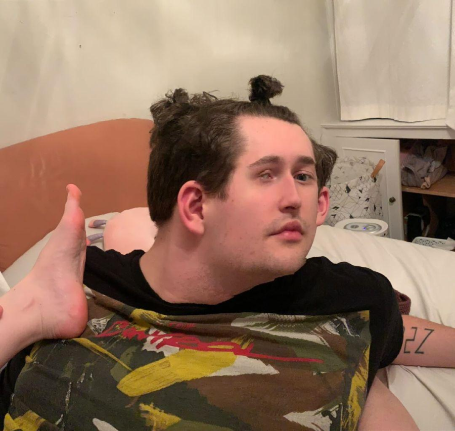 A man lying on a bed with his hair in two small buns. One of his legs is lifted up so that his foot is resting on his shoulder, creating an unusual and humorous pose. He is looking slightly to the side.