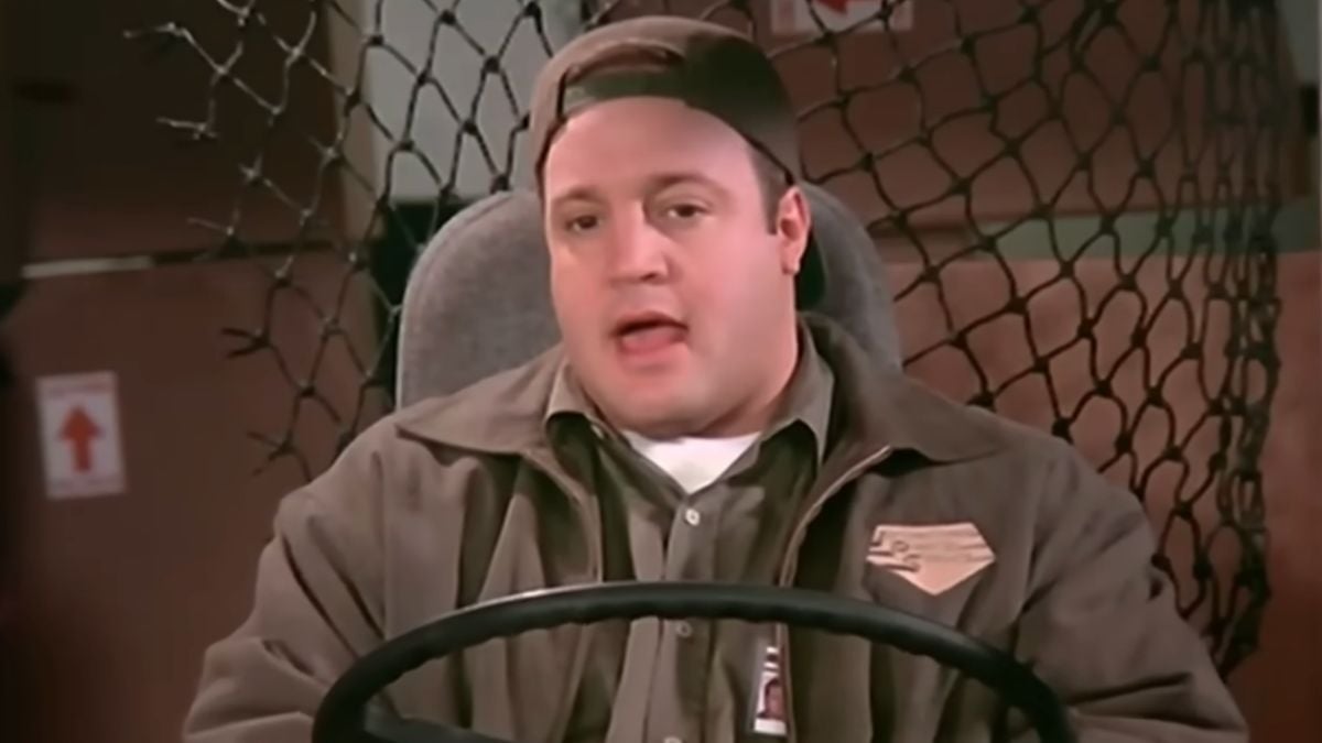 A man in a brown delivery uniform and cap sits behind the wheel of a vehicle with a mesh divider and cardboard boxes in the background.