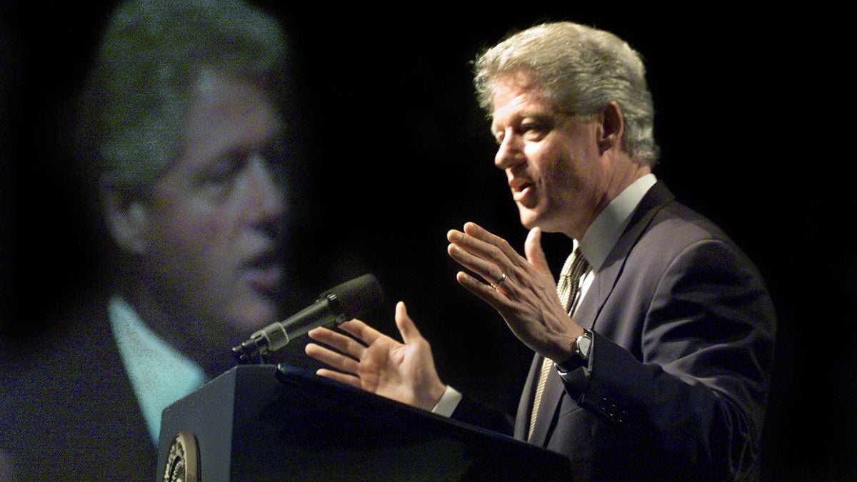A man in a suit speaks at a podium with a microphone, gesturing with his hands. His large, blurred image is projected on a screen behind him against a dark background.