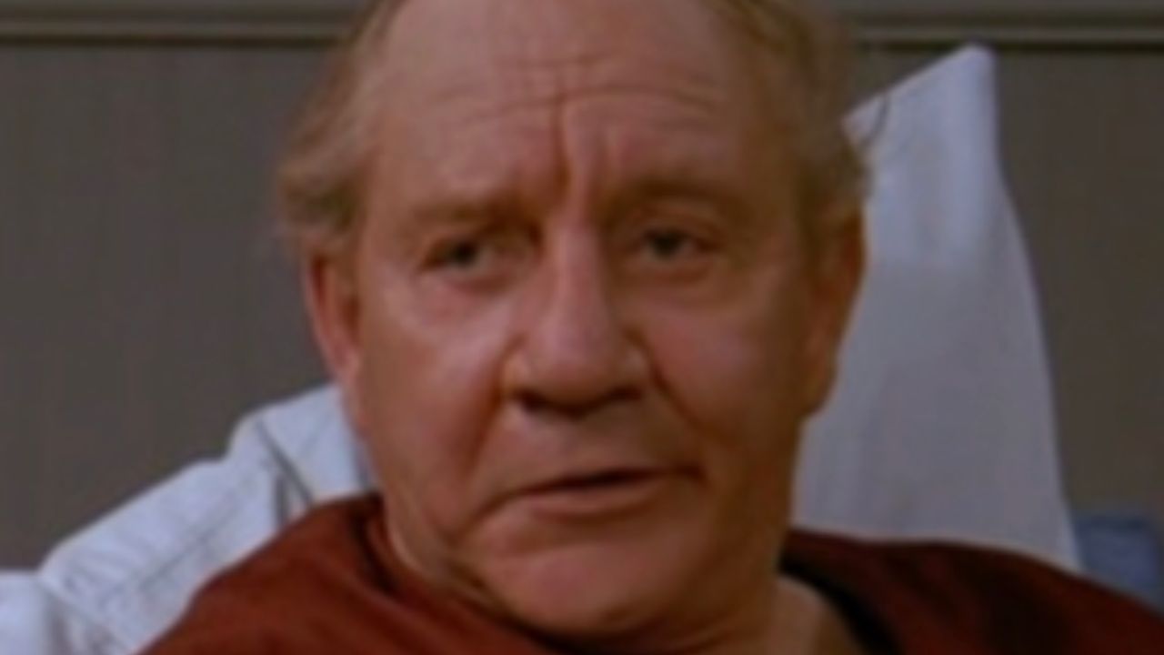 An older man with light hair sits up in a hospital bed, wearing a maroon shirt, with a neutral expression. The background is out of focus.