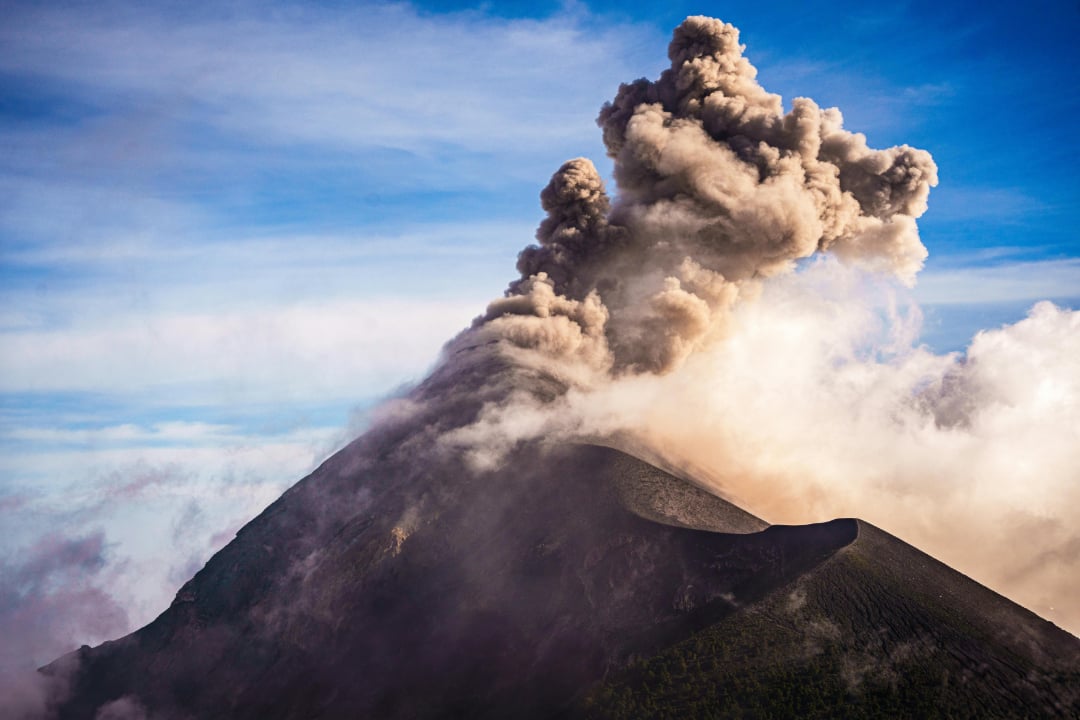 A dark volcano erupts, sending thick clouds of gray ash and smoke high into a blue sky, with the slopes partly covered in ash and vapor.