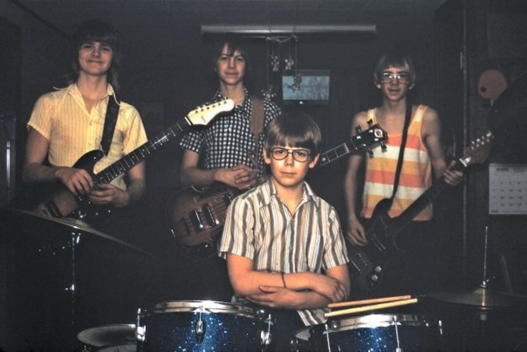Four boys in a dimly lit room pose with musical instruments. Three stand in back with electric guitars and a bass, while one sits in front at a drum set, arms crossed, all wearing casual striped shirts.