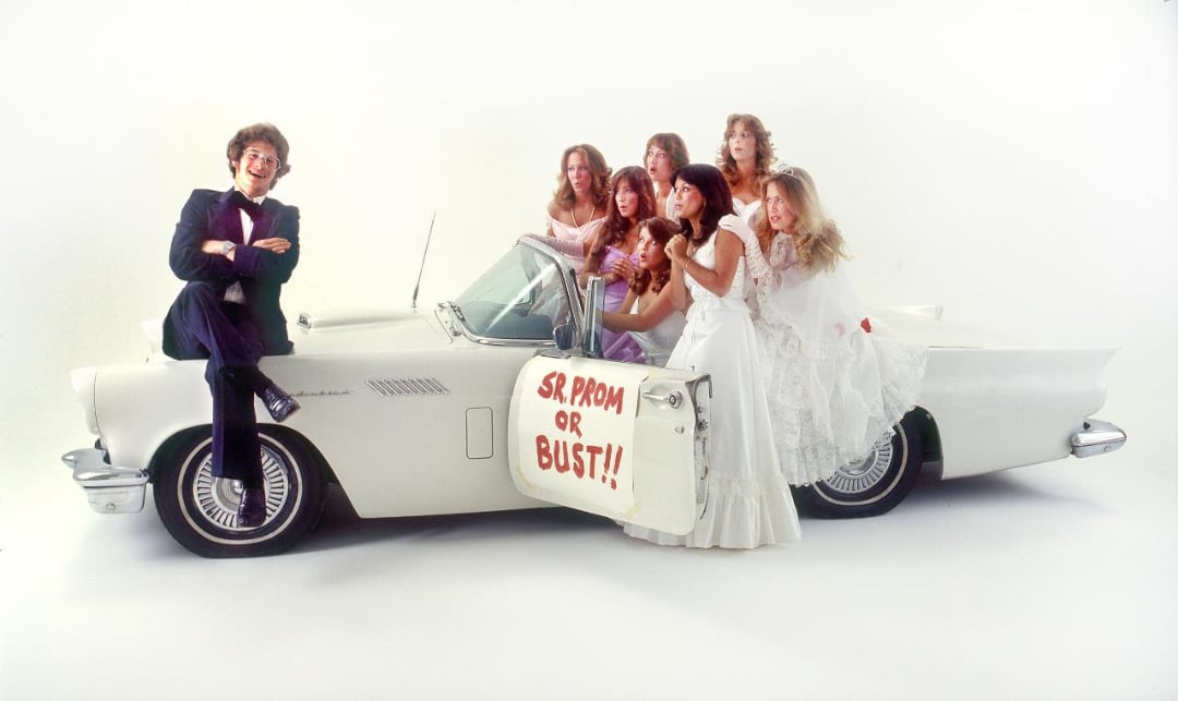 A group of teens in formal prom attire pose with a white vintage convertible. Six girls in white dresses sit inside, while a boy in a suit sits on the hood. A sign on the door reads “SR. PROM OR BUST!!” The background is plain white.