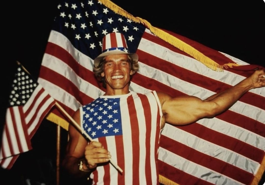 A smiling man wearing a sleeveless shirt and top hat patterned like the American flag holds two U.S. flags, with a large flag draped behind him. The background is dark.