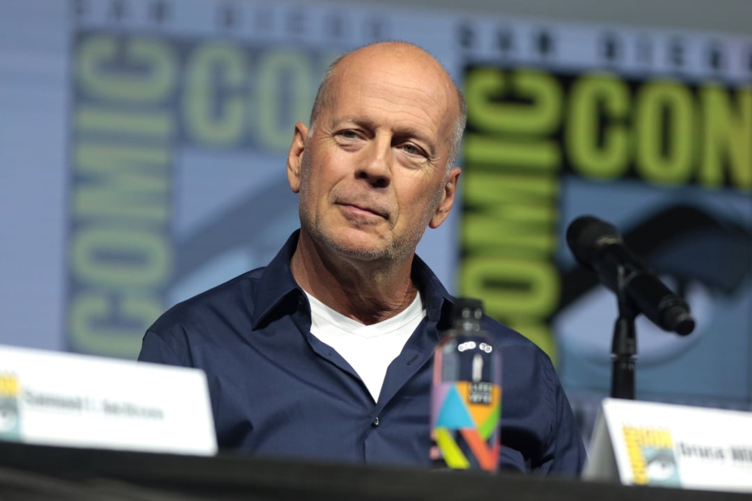 A man with a bald head wearing a dark blue shirt and white undershirt sits at a panel table with a microphone and a colorful water bottle. A Comic-Con banner is visible in the background.