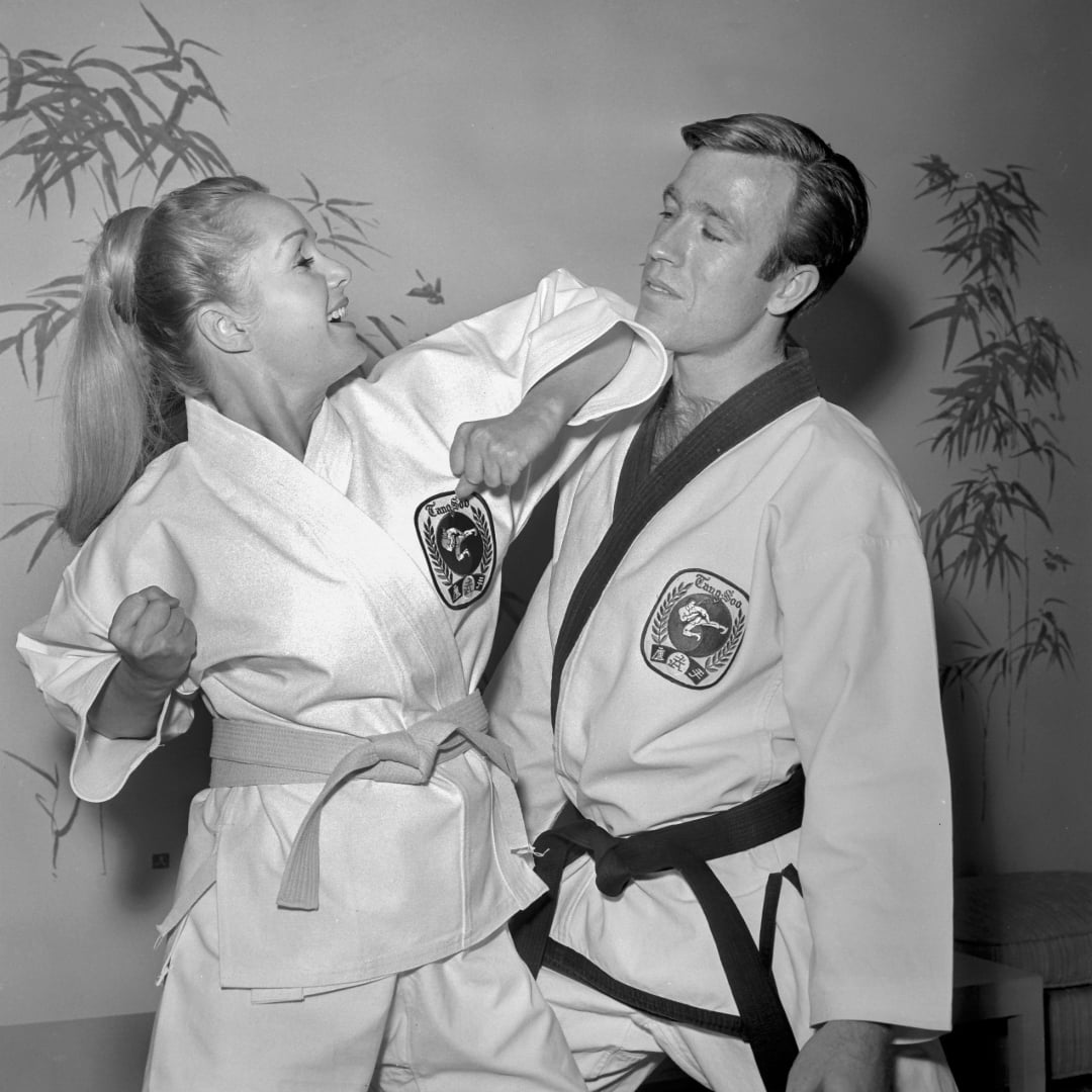 A woman in a martial arts uniform demonstrates a self-defense move, striking a man in a similar uniform. Both have focused expressions, and bamboo decorations are visible in the background.