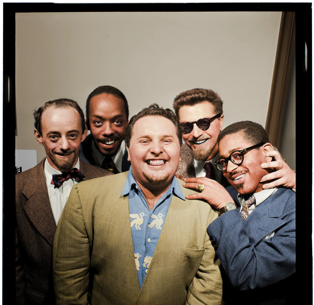 Five men in vintage suits pose closely together, smiling and laughing for the camera. The man in the center wears a light-colored jacket and patterned shirt; two men wear glasses. The mood is friendly and cheerful.
