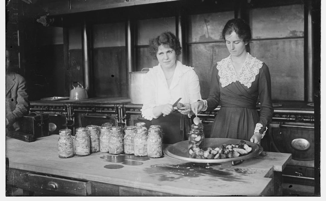Two women stand at a kitchen table, filling glass jars with cut vegetables from a large tray. Several jars, some already filled, are lined up on the table. The kitchen has a stove and utensils in the background.