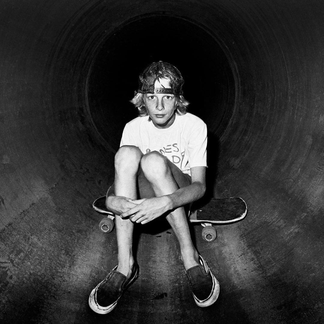 A young person with long hair and a headband sits cross-legged on a skateboard inside a large concrete pipe, wearing a T-shirt and slip-on shoes, looking directly at the camera in black and white.