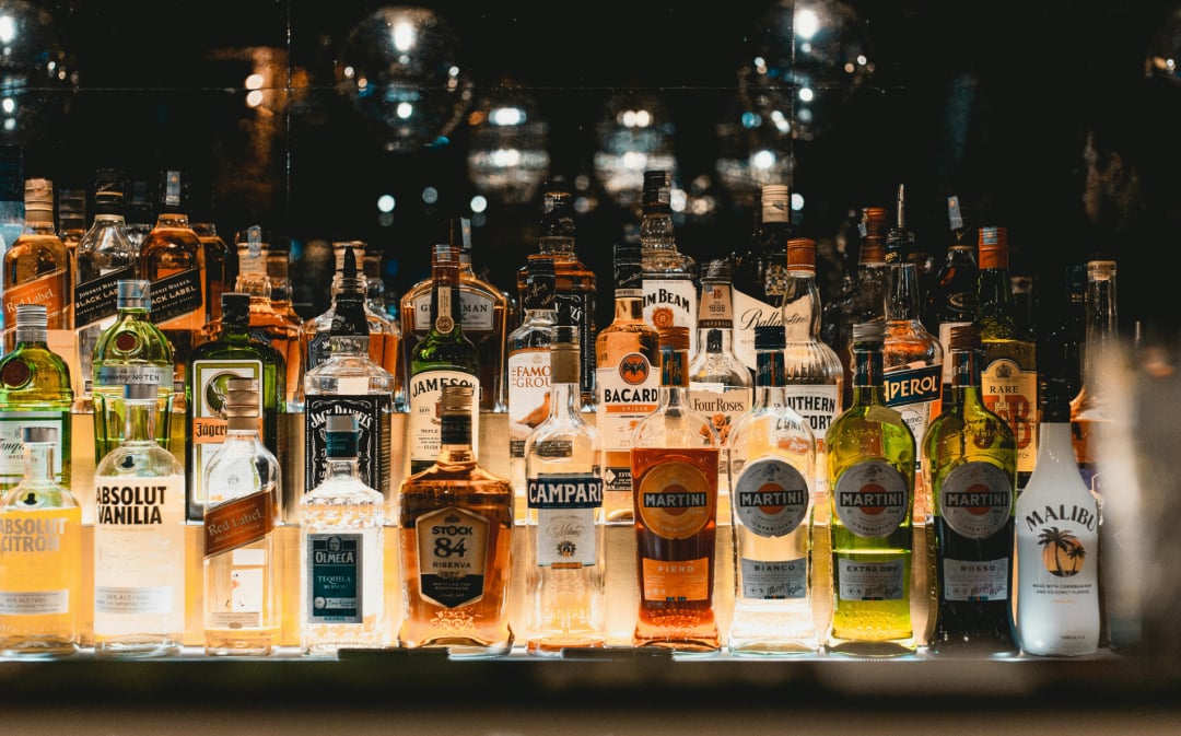 A variety of liquor bottles, including whiskey, vodka, rum, and liqueurs, are arranged on a bar shelf with warm lighting reflecting off the glass. Brands such as Absolut, Bacardi, Campari, and Martini are visible.