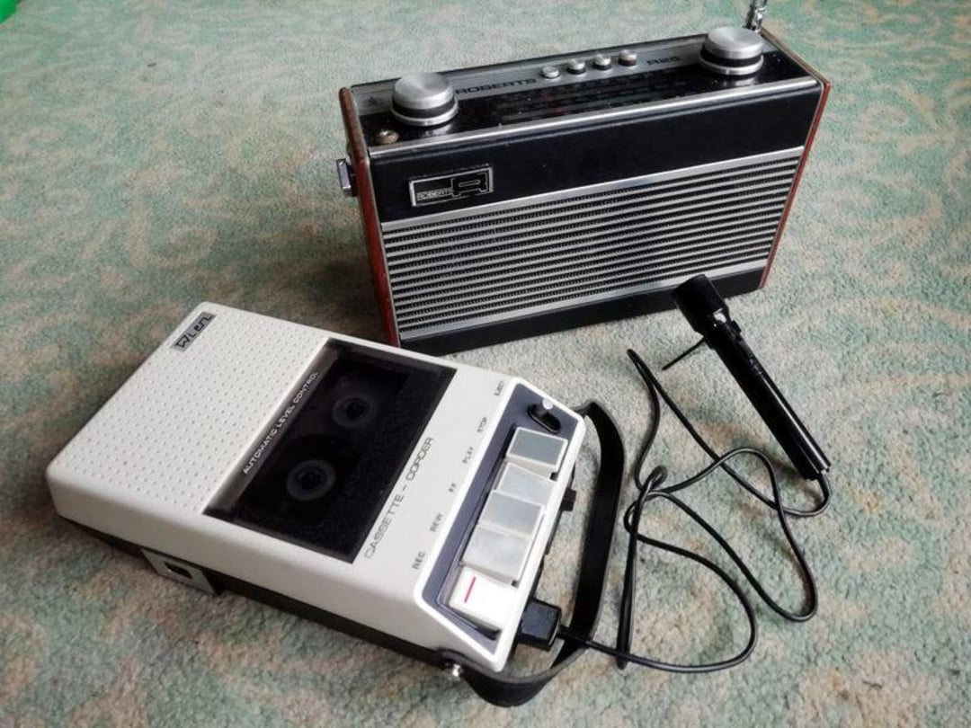 A vintage radio with a metal grille stands upright next to a white cassette recorder with large buttons and a connected black microphone, all placed on a light patterned carpet.