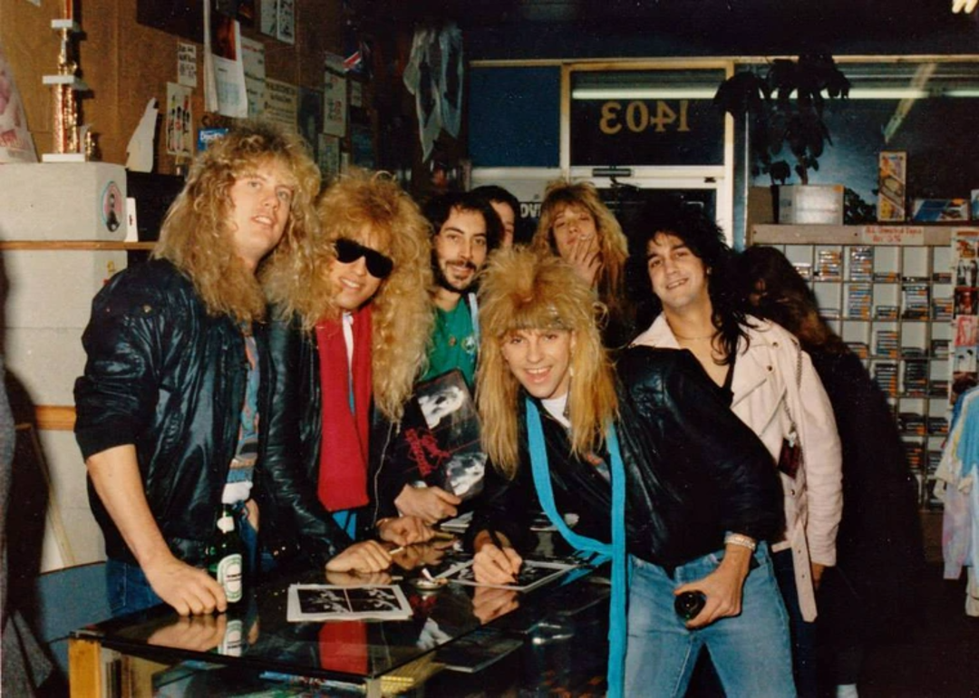 A group of six people from different schools with big 1980s hairstyles and leather jackets smile and pose for a photo inside a music store, some holding photographs and drinks, with shelves of cassette tapes in the background.