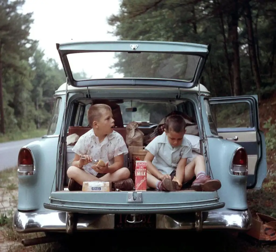 Two children sit in the open trunk of a packed blue station wagon, eating snacks. A carton of milk and bags are beside them. The car is parked by a wooded roadside on a sunny day.