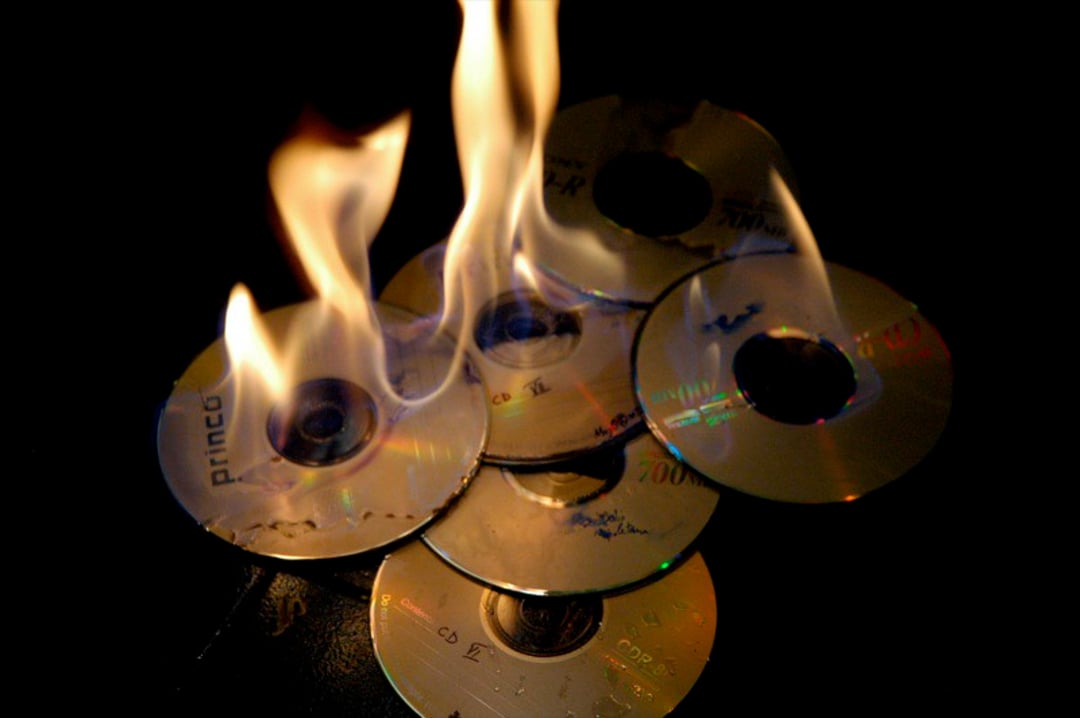 A stack of six CDs is burning, with bright orange flames rising above them against a dark background.