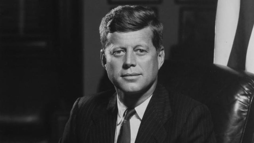 A man in a dark suit and tie sits in a chair, looking directly at the camera. There is an American flag partially visible behind him, and the background is dimly lit.