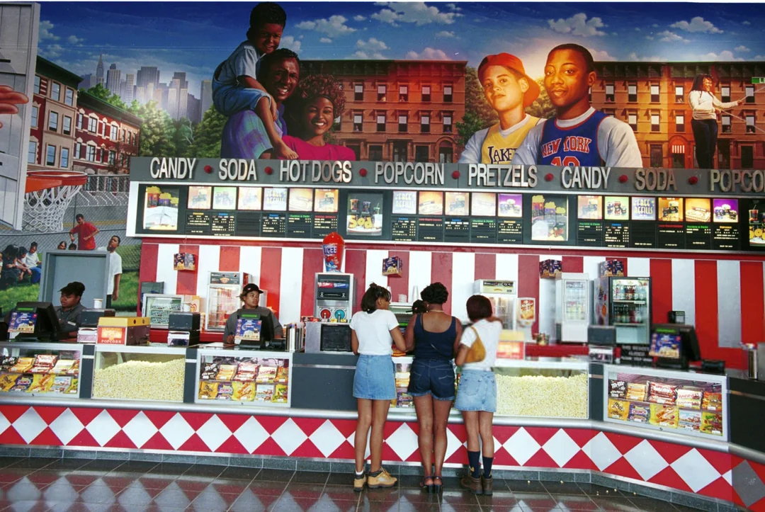 Three people stand at a movie theater concession counter with a colorful mural of city scenes and people above. The counter displays popcorn, snacks, and menu boards.
