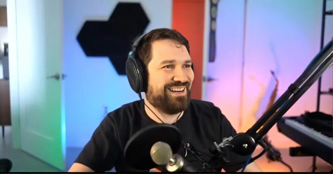 A man with headphones and a beard smiles while sitting at a desk with a microphone in front of him. The room has colorful lighting, a keyboard, and soundproofing panels on the wall.