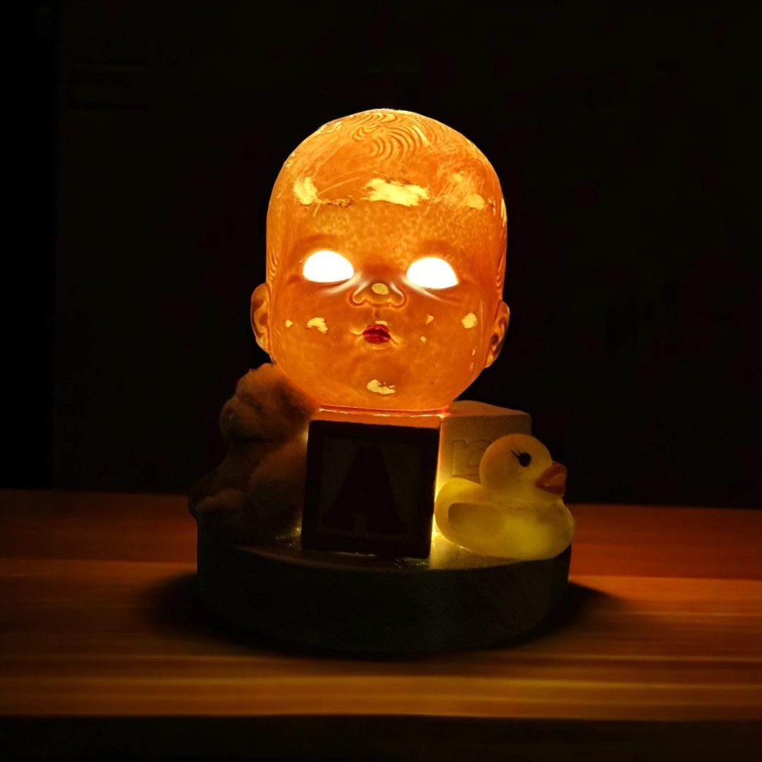 A glowing lamp shaped like a doll's head with illuminated eyes, placed on a table beside a yellow rubber duck, a small teddy bear, and a red letter block with "A." The scene is dimly lit and eerie.
