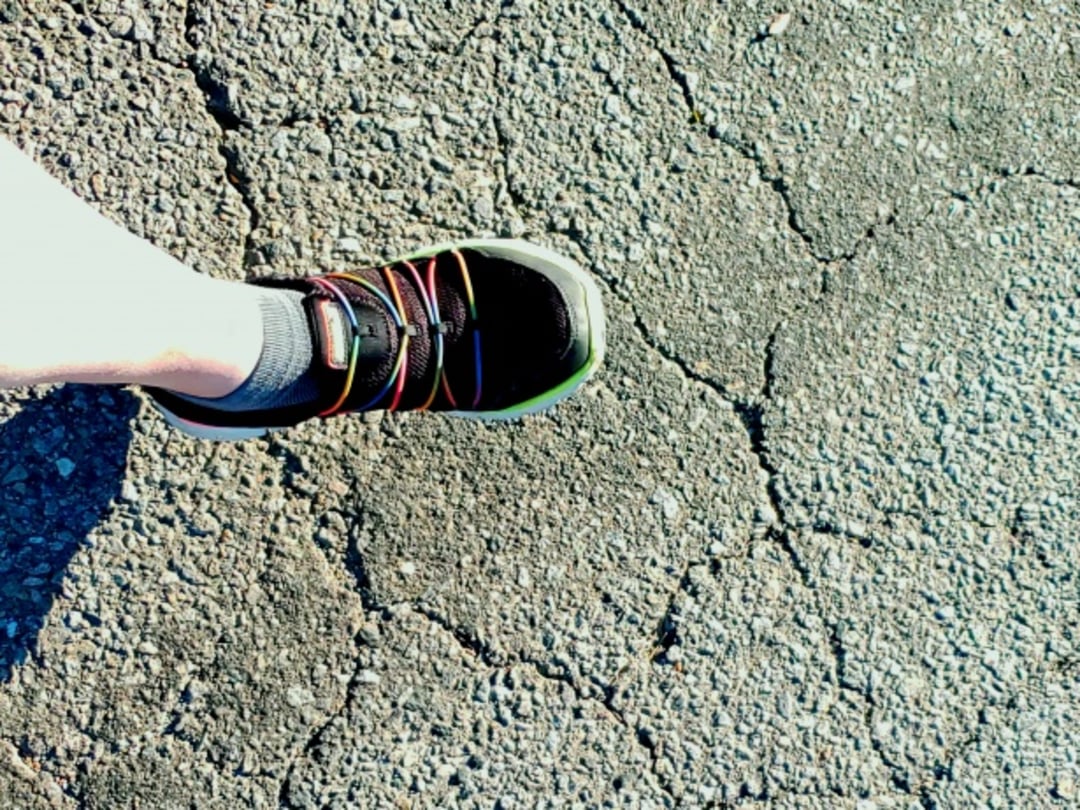 A person wearing a black sneaker with colorful laces and a gray sock stands on sunlit, cracked asphalt pavement.