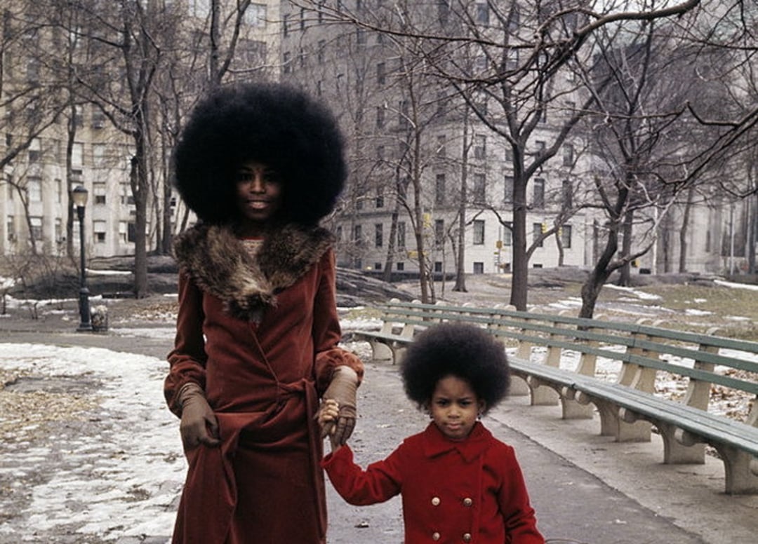 A woman and a young girl, both with natural afros and wearing red coats, stand hand-in-hand on a path in a city park with leafless trees and patches of snow on the ground.