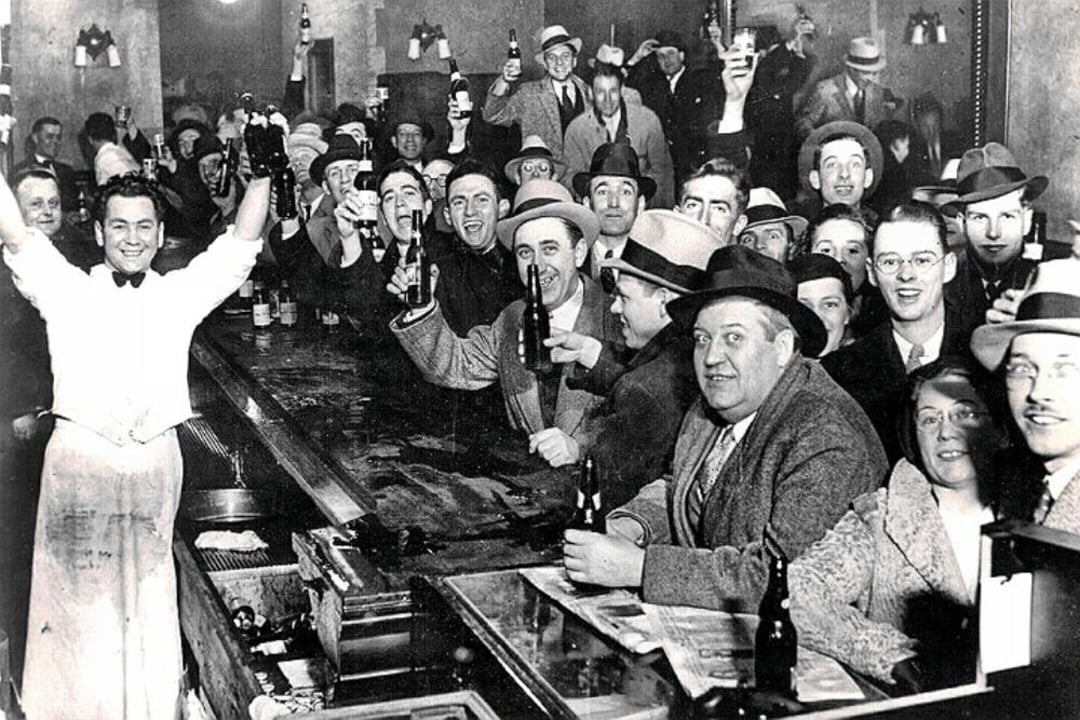 A crowded bar scene from the early 20th century shows smiling patrons in suits and hats raising bottles, with a cheerful bartender standing behind the bar, arms raised in celebration.