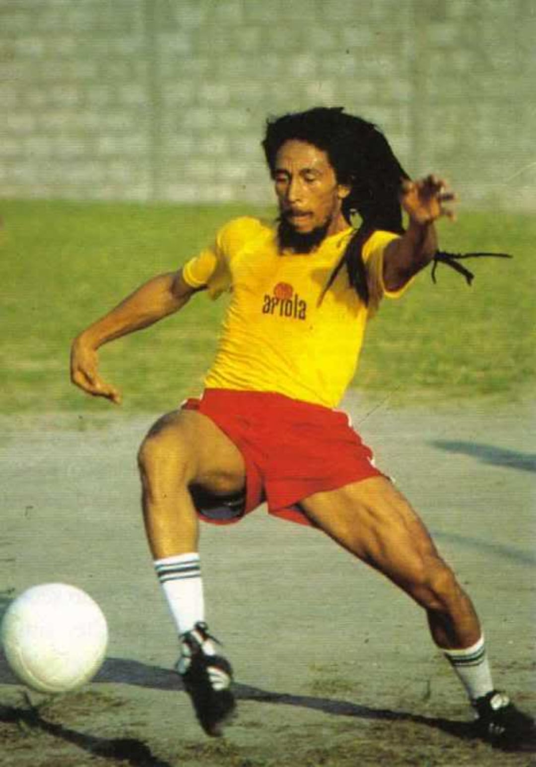 A man with long dreadlocks wearing a yellow shirt and red shorts plays soccer outdoors, mid-action as he prepares to kick a white ball on a grass and dirt field.