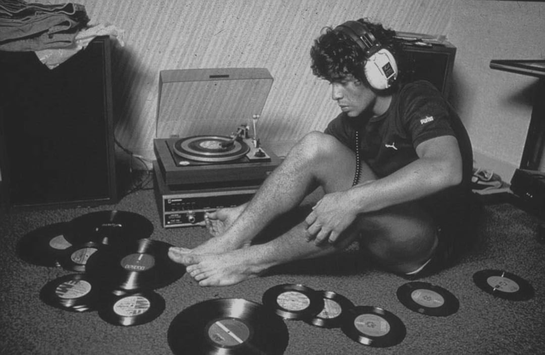 A young man wearing headphones sits on the floor next to a record player, surrounded by vinyl records, appearing deep in thought as he listens to music.