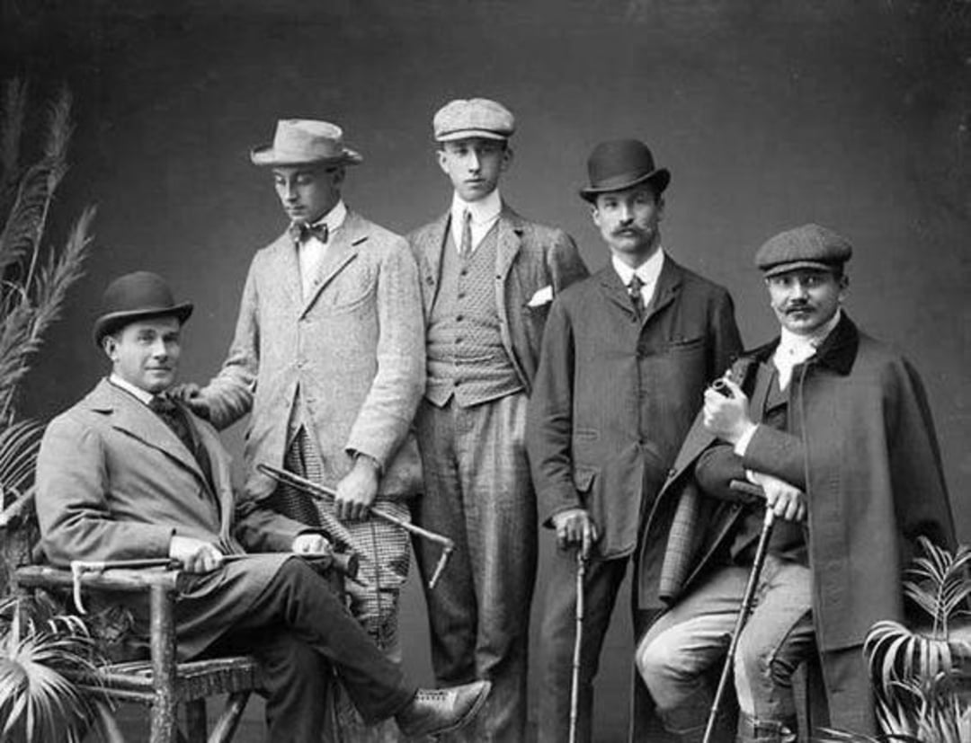 Five men in early 20th-century attire pose for a formal portrait; three stand while two are seated. They wear suits, vests, bowler hats, and flat caps, with canes and props, against a studio backdrop with plants.