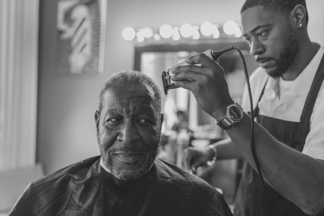 A barber trims an older man's hair with clippers in a bright, modern barbershop. The man sits in a chair wearing a cape, while the barber concentrates on his work. The image is in black and white.