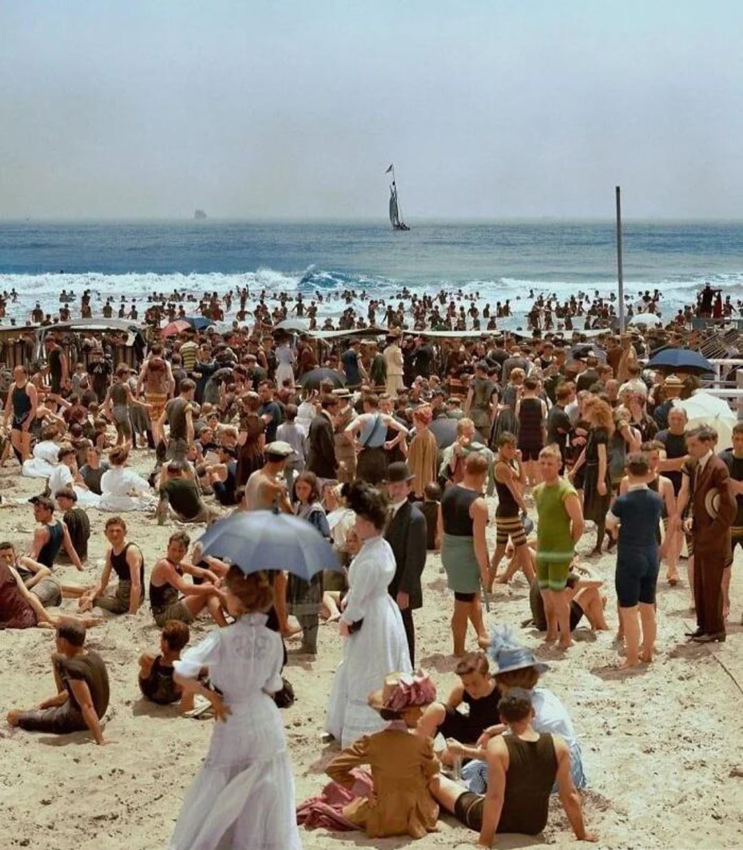 A crowded beach scene from the early 1900s shows people in vintage swimsuits and dresses relaxing, socializing, and enjoying the sunny day by the ocean, with a sailboat visible in the distance on the water.
