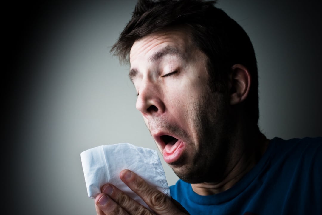 A man with dark hair sneezes into a white tissue, eyes closed and mouth open, against a dark background.