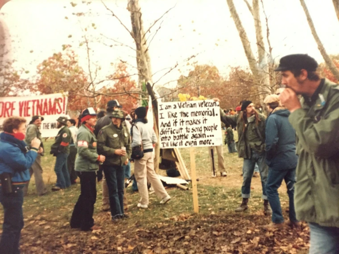 A group of people stand outside among trees holding signs. One sign reads, “I am a Vietnam veteran. I like the memorial. And if it makes it difficult to send people into battle again, I like it even more.”