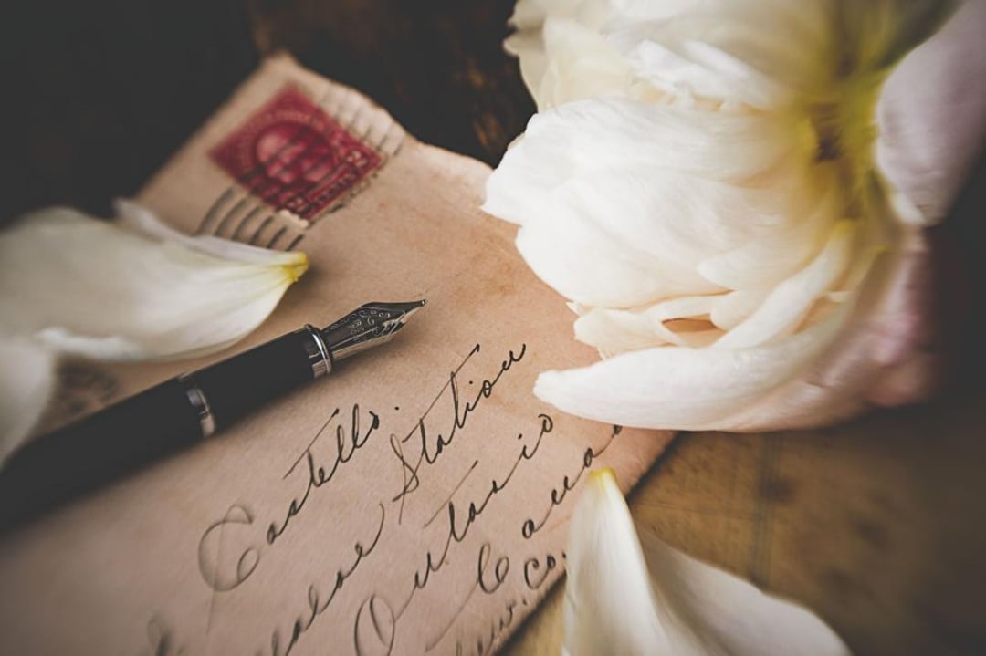 A vintage envelope with handwritten calligraphy, a fountain pen, and white flower petals rests on a surface, creating a nostalgic, romantic atmosphere.