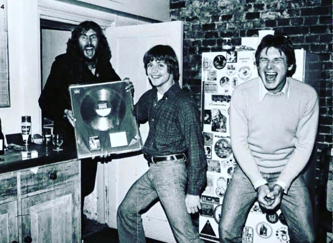 Three young men stand in a kitchen, smiling and celebrating. One holds a framed record award, while the others laugh. The room has a brick wall and a fridge covered in stickers. The image is in black and white.