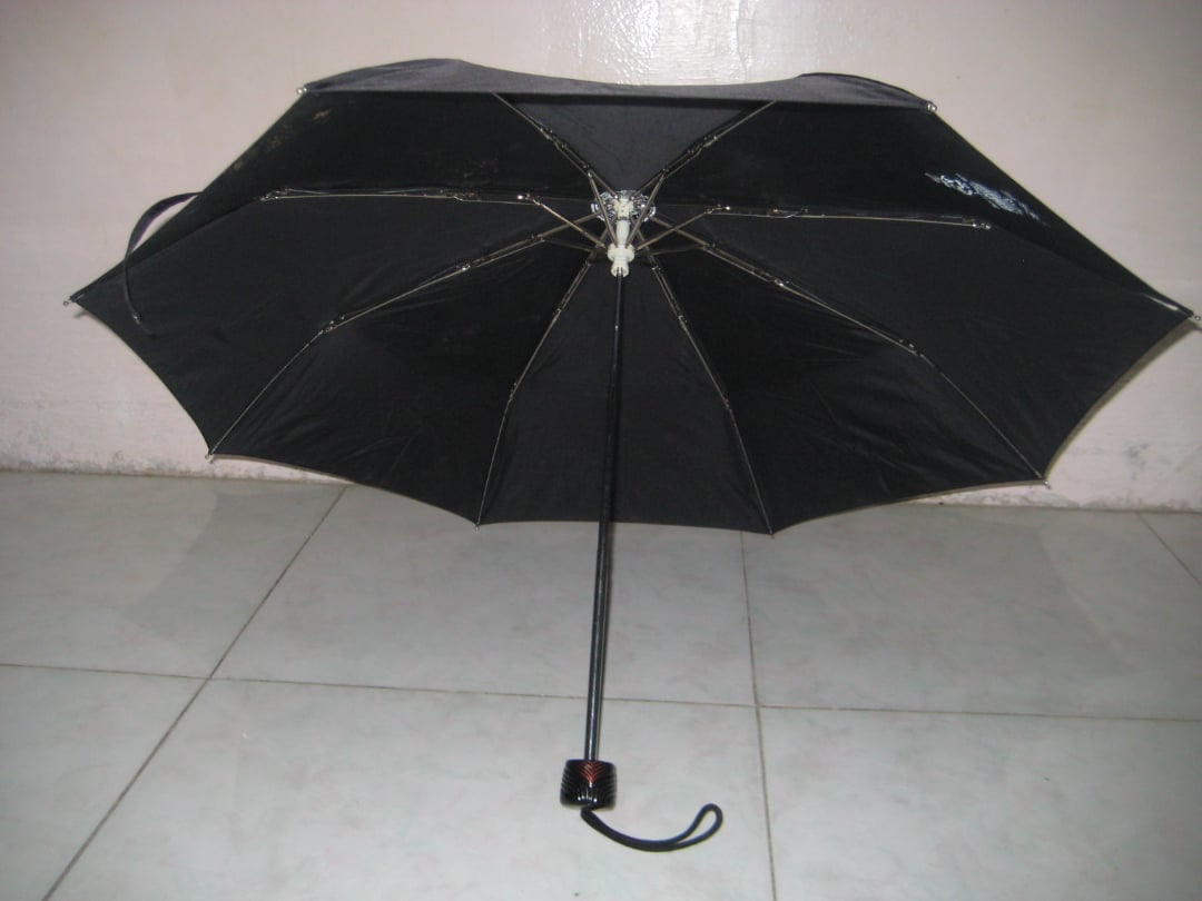 A black umbrella is open and resting on a tiled floor, leaning against a light-colored wall. The handle is curved and the umbrella's canopy is fully extended.