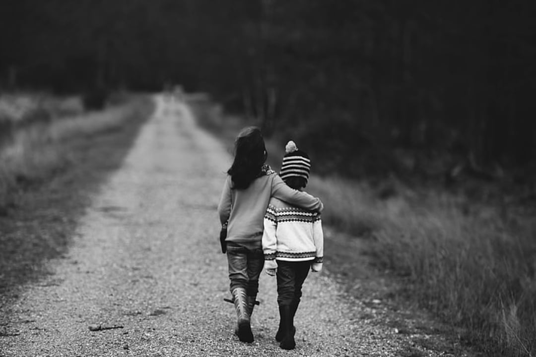 Two children walk down a gravel path, one with their arm around the other's shoulders. The scene is in black and white and the background shows trees and grass along the path.