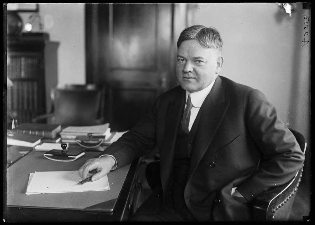A man in a suit sits at a desk holding a pen over a blank sheet of paper, looking toward the camera. There are books, papers, and office items on the desk, with a bookshelf and a wooden door in the background.