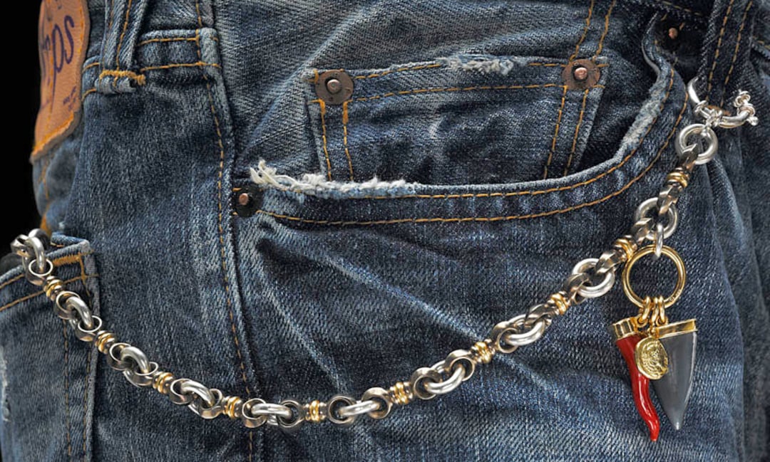 A close-up of blue denim jeans with a decorative silver and gold chain attached to a belt loop and front pocket, featuring two charms—a red horn and a small black horn with a gold emblem.