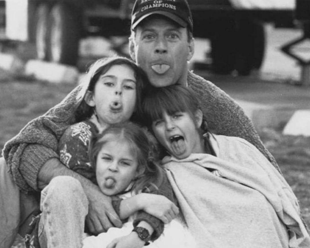 A man wearing a cap and three young girls sit close together outdoors, all making funny faces and sticking out their tongues for the camera. The photo is in black and white.