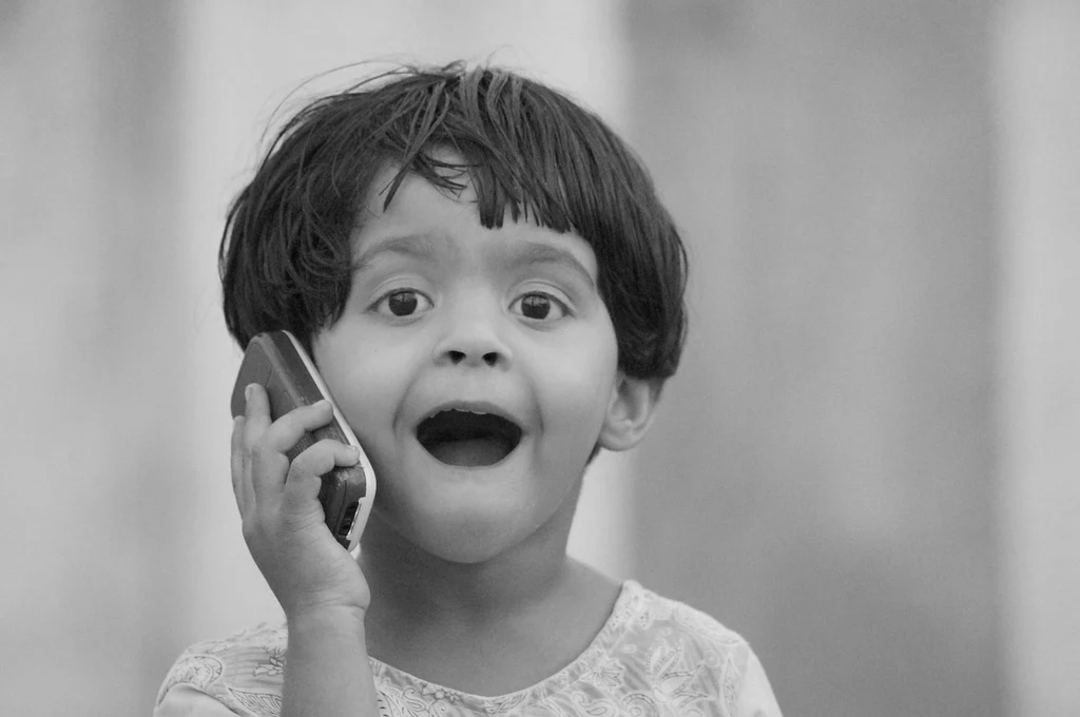 A young child with short hair holds a phone to their ear, looking surprised or excited, with their mouth open and eyes wide. The image is in black and white.