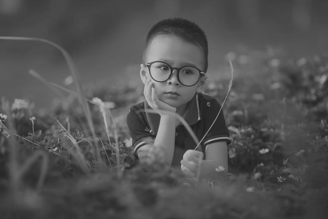 A young boy wearing glasses lies in a field of flowers, resting his chin on his hand and looking thoughtful. The image is in black and white.