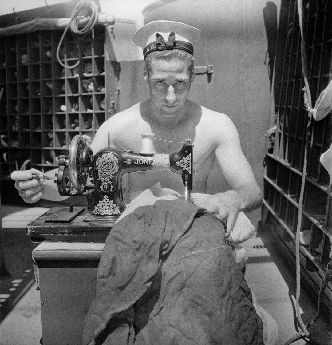 A shirtless sailor wearing a hat sits at a sewing machine, stitching fabric on a ship. Storage cubbies and metal structures are visible in the background.