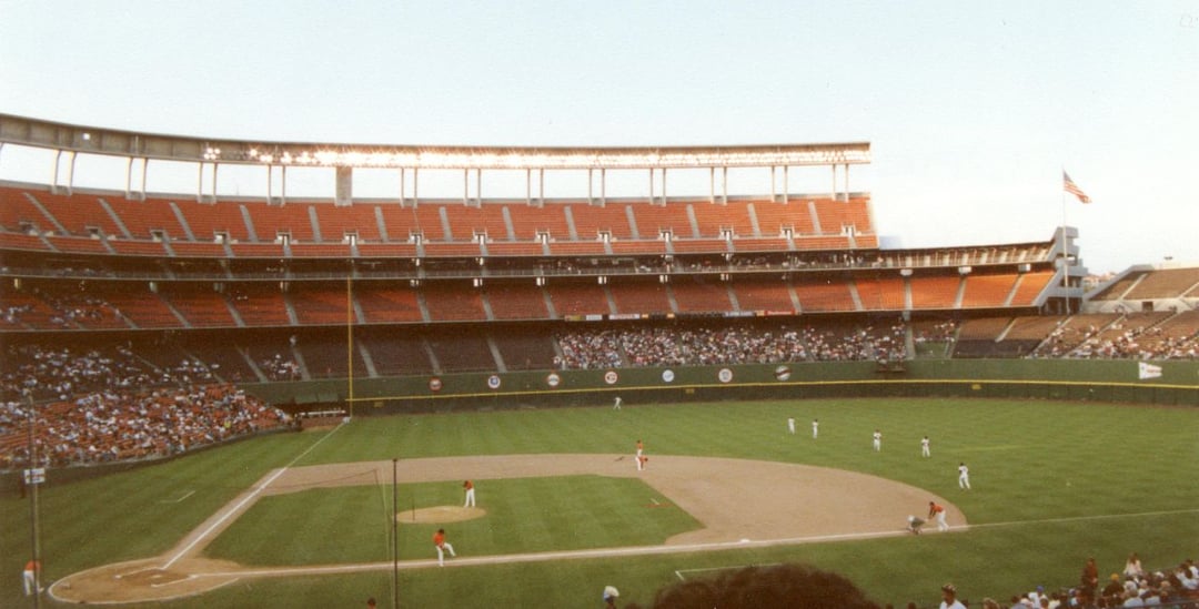 A baseball stadium with mostly empty orange seats; a game is in progress on the field with players visible and a few spectators scattered in the stands. An American flag waves in the background.