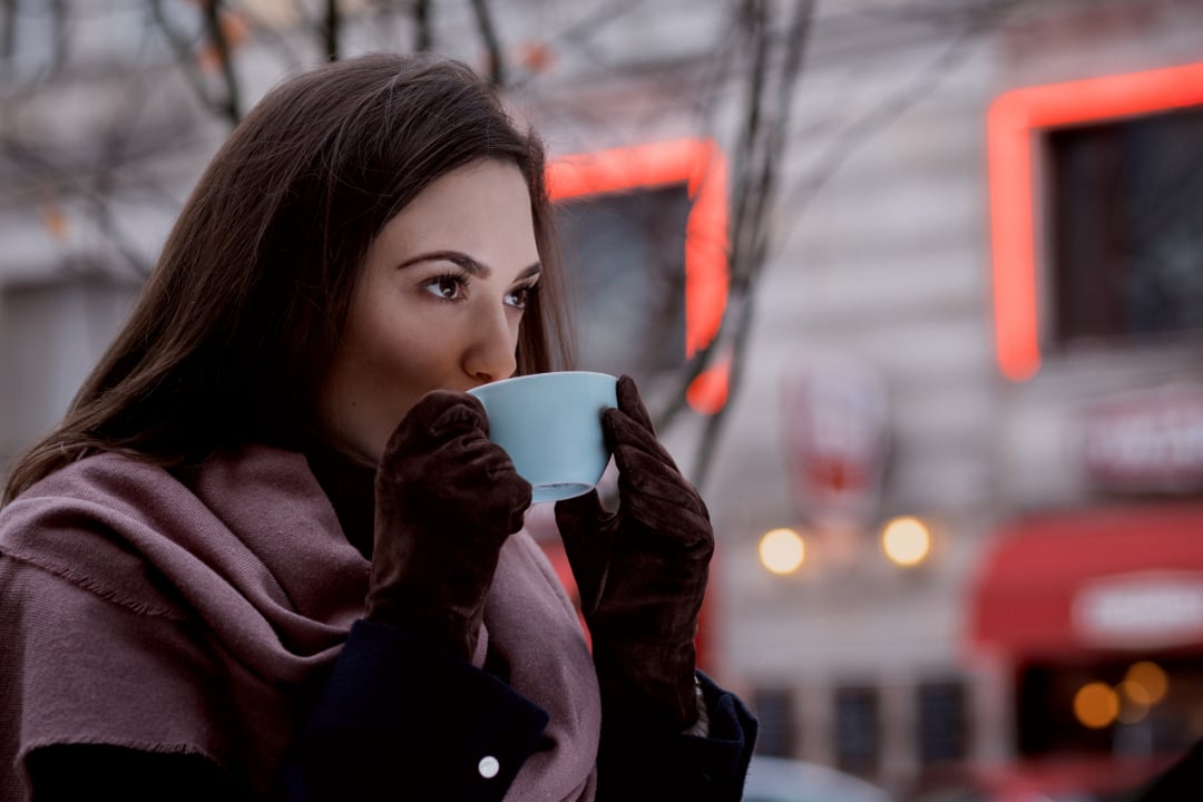 A woman with long brown hair, wearing a pink scarf and brown gloves, holds a light blue cup close to her face and drinks, outdoors in a city with blurred red and white signage in the background.