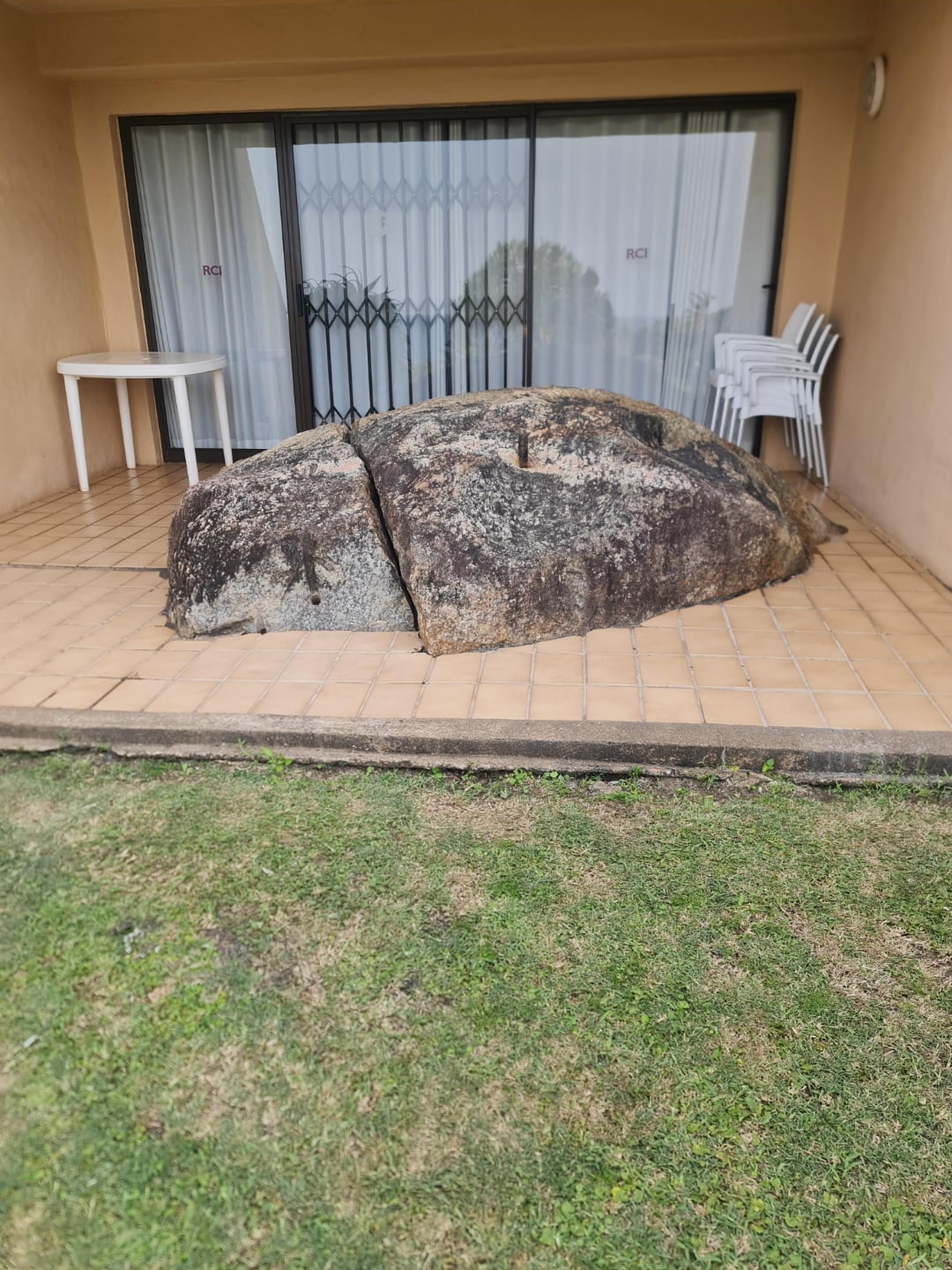 A large rock sits embedded in the tiled patio of a building, partially sheltered by the roof. White plastic chairs and a table are stacked on one side near sliding glass doors with security bars. Grass borders the patio.