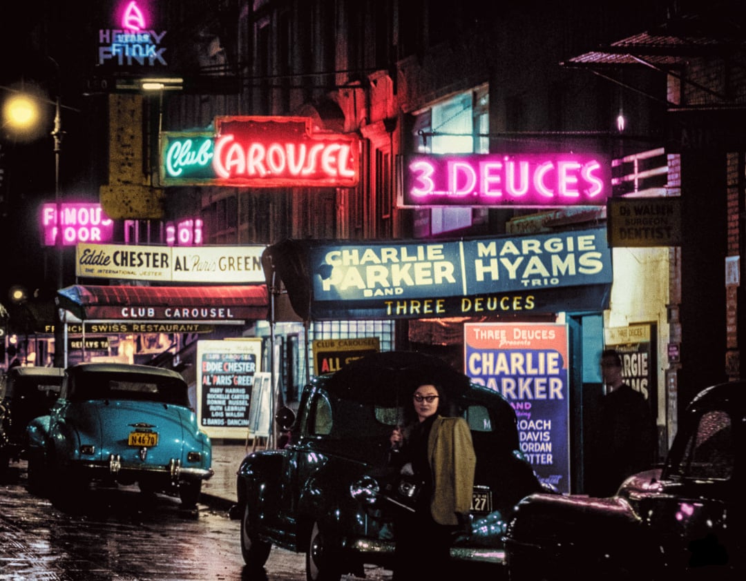 A woman stands on a wet city street at night, surrounded by vintage cars and illuminated by colorful neon signs advertising jazz clubs like “Club Carousel” and “Three Deuces” featuring Charlie Parker.