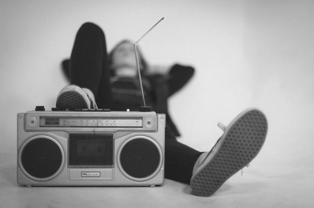 A person sits relaxed with their legs stretched out, one foot resting on a large vintage boombox radio. The image is black and white, and the person is out of focus in the background.