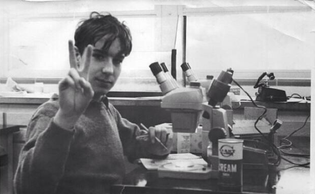 A young person sitting at a desk with microscopes holds up a peace sign. The desk has scientific equipment and a can labeled "Dream." The background shows large windows and a classroom or laboratory setting.